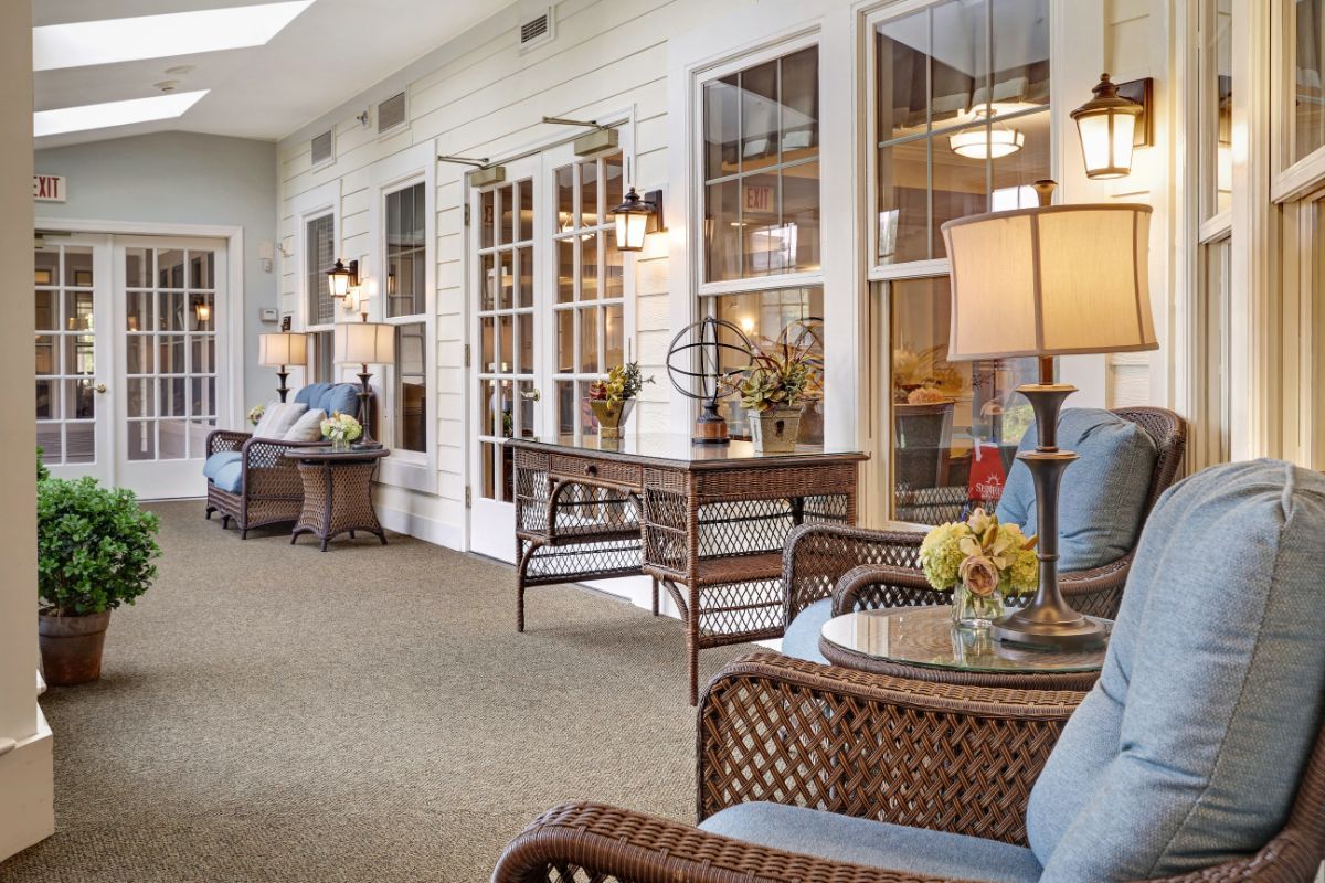 A cozy interior space featuring wicker chairs and tables adorned with lamps and flowers, set against large windows.