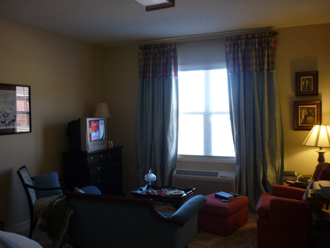 A cozy living room with a television, sofa, and chairs near a curtained window.