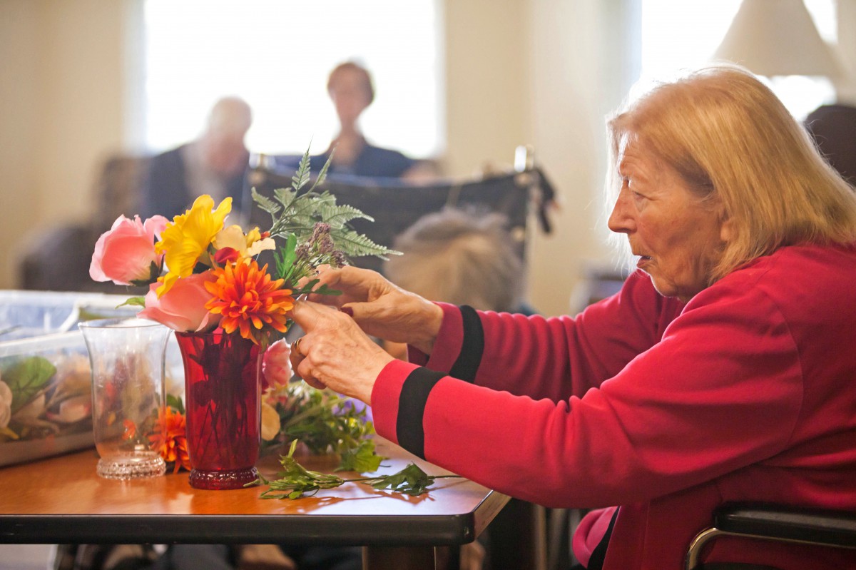 An elderly woman in a red sweater carefully arranges flowers in a vase indoors.