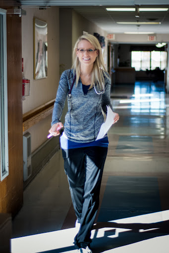 A nurse in scrubs walks through a hospital corridor, holding paperwork and wearing a stethoscope.