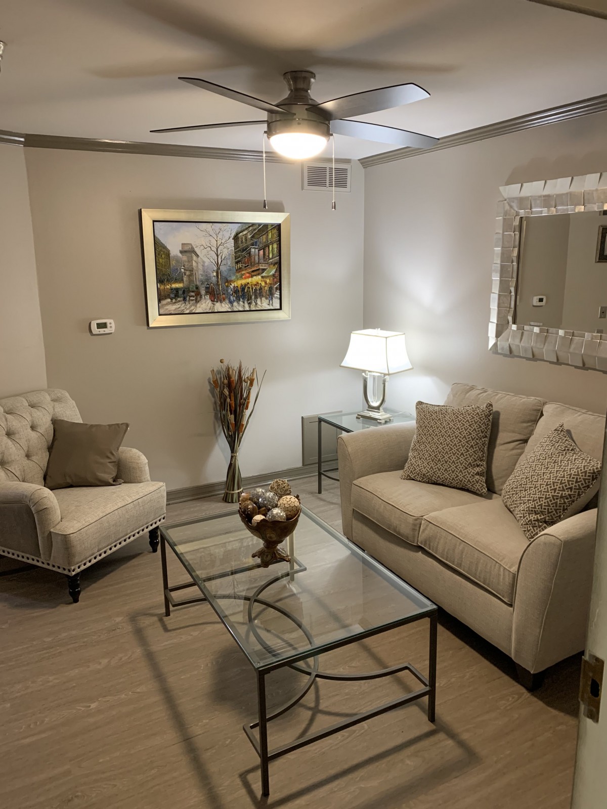A cozy living room with elegant furniture and decor featuring a sofa, armchair, glass coffee table, lamp, wall art, and a mirror.