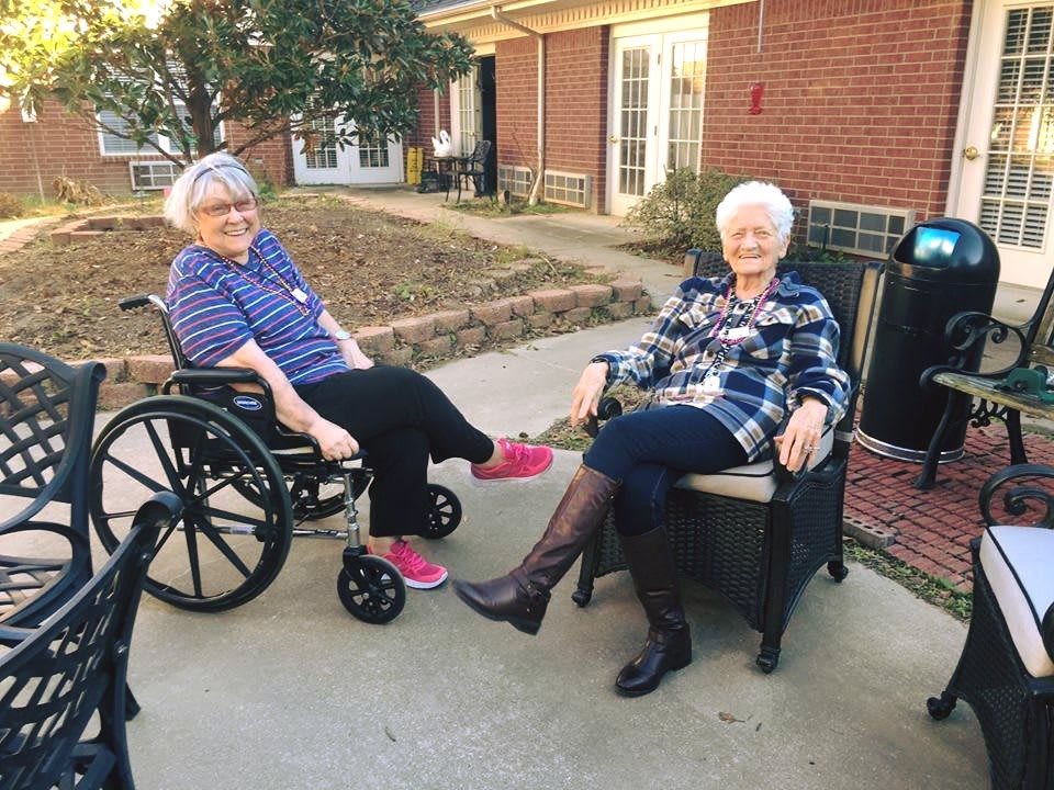 Two elderly women enjoying a sunny day outdoors, one in a wheelchair and the other seated in a chair by a brick building.