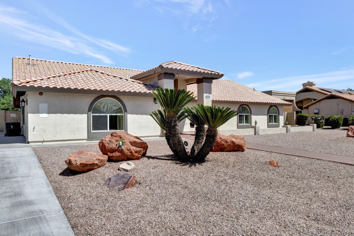 A stucco house with a tile roof, featuring desert landscaping with palm plants and rocks.