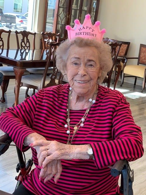 An elderly woman wearing a pink birthday tiara and a red striped shirt, seated indoors.