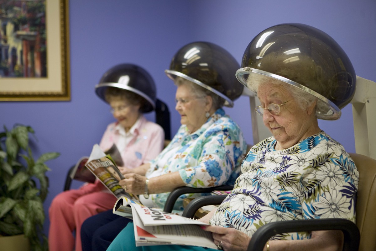 Elderly women sitting under hair dryers and reading in a beauty salon.