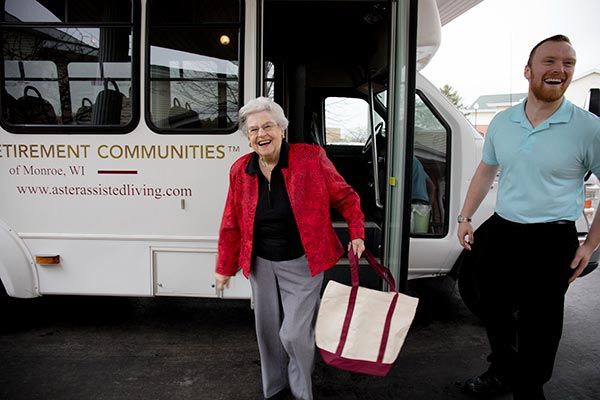 An elderly woman in a red jacket smiles as she steps off a bus carrying a bag.