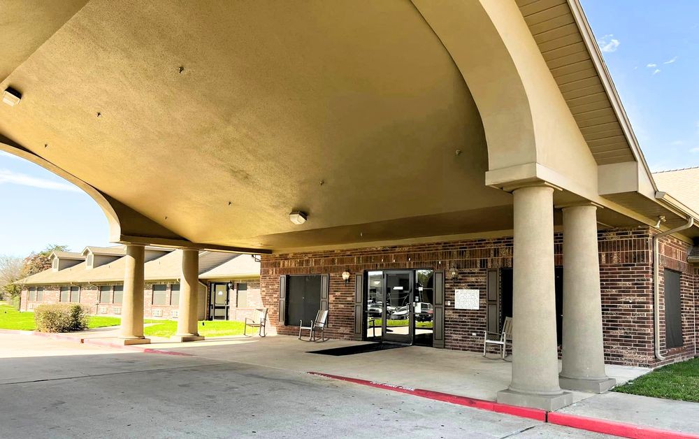 The entrance of a building featuring a covered walkway supported by columns, leading to glass doors.