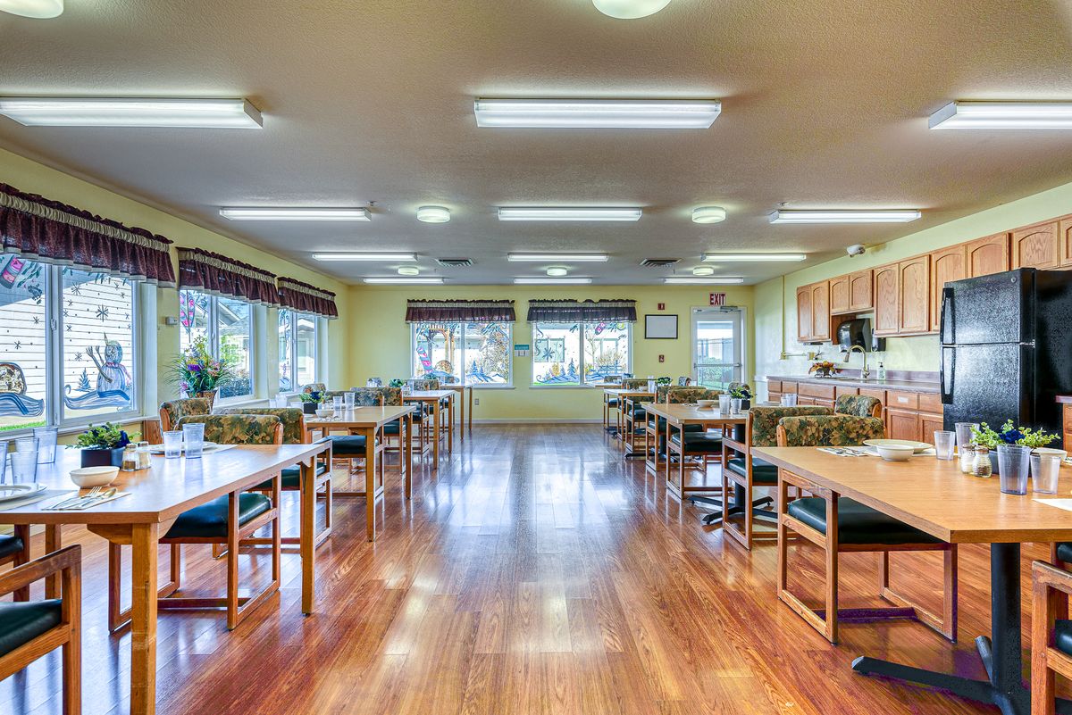 A spacious dining area with wooden flooring and ample natural light, featuring multiple tables and a kitchen area.