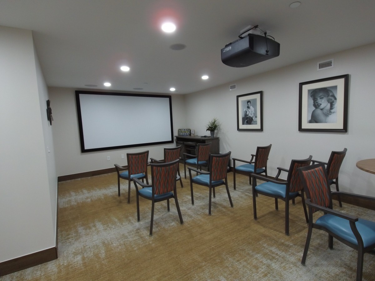 A small home theater setup with several chairs facing a large screen and projector, featuring wooden flooring and framed pictures on the walls.