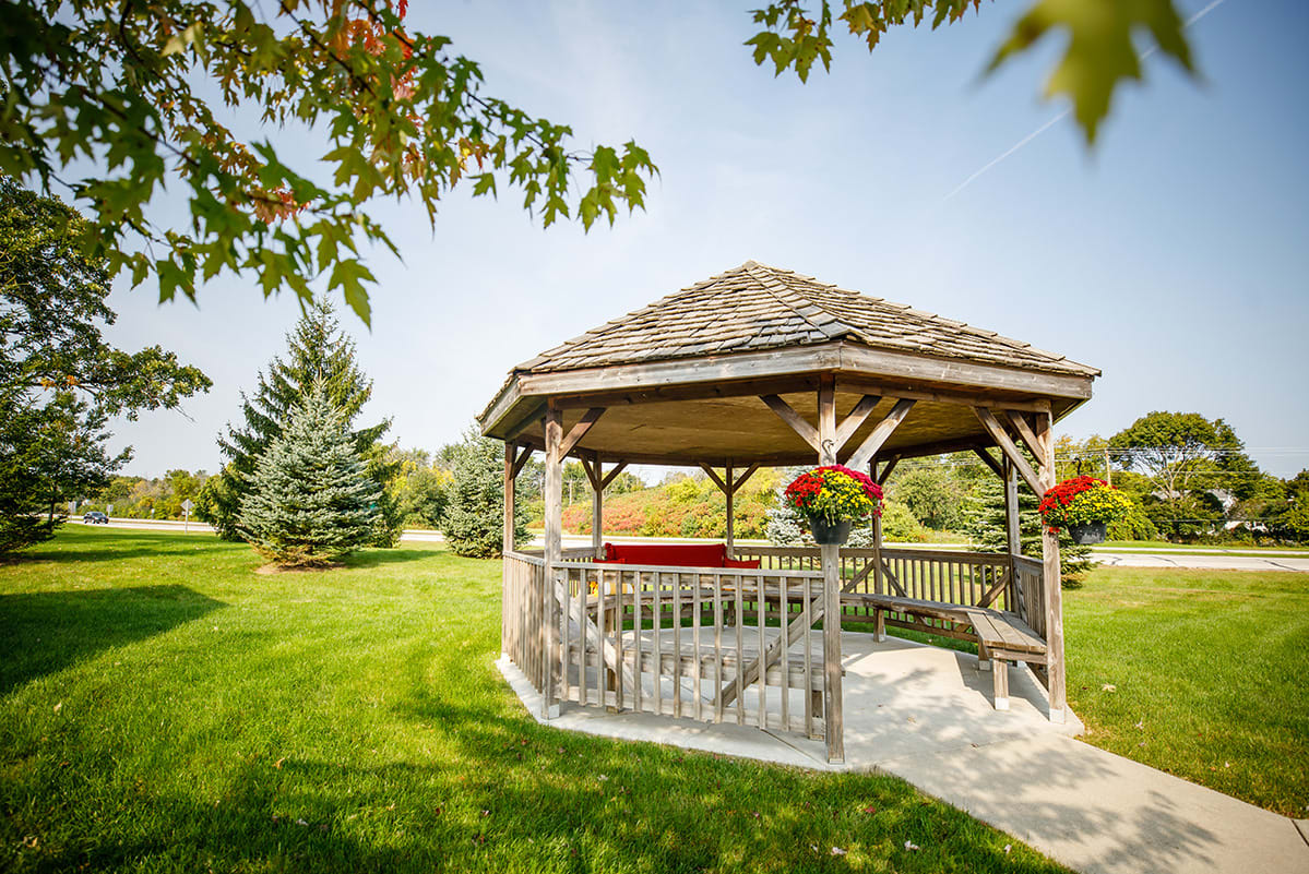 A serene gazebo in a lush green park, surrounded by trees and flowers under a clear blue sky.