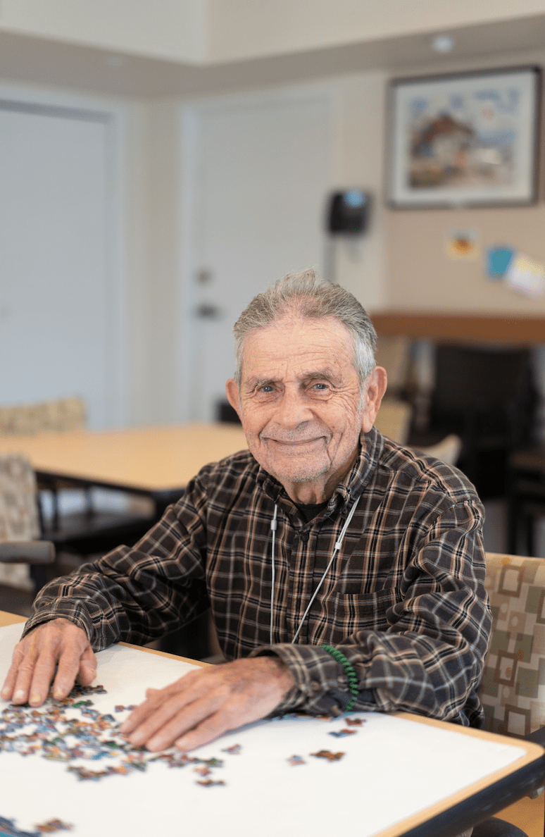An elderly man in a plaid shirt smiling while sitting at a table indoors.