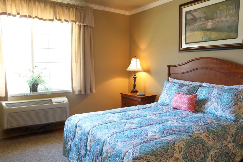 A cozy bedroom featuring a bed with a blue patterned quilt, a bedside lamp, and a window with curtains.