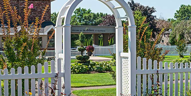 A charming garden view through a white archway with vibrant green plants and a picket fence.