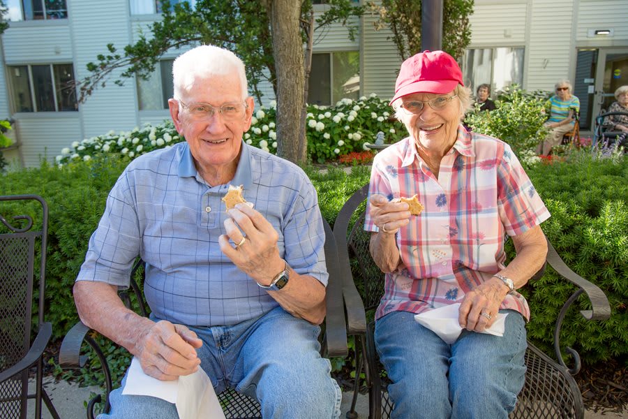 Two elderly people enjoying a snack while sitting on a park bench.