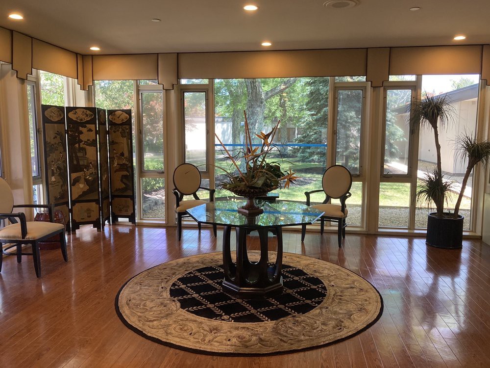 A stylish living room with a glass table centerpiece, surrounded by chairs and large windows allowing ample natural light.
