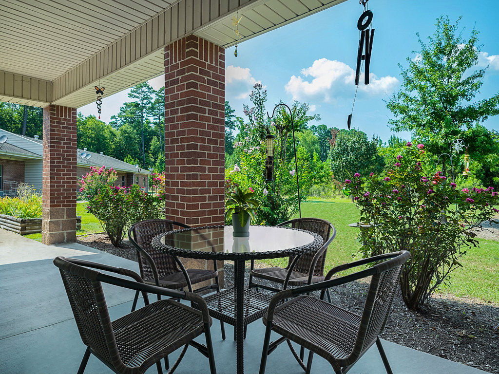 A cozy patio setup with a glass-top table and chairs, surrounded by greenery and brick pillars, offering a serene backyard view.
