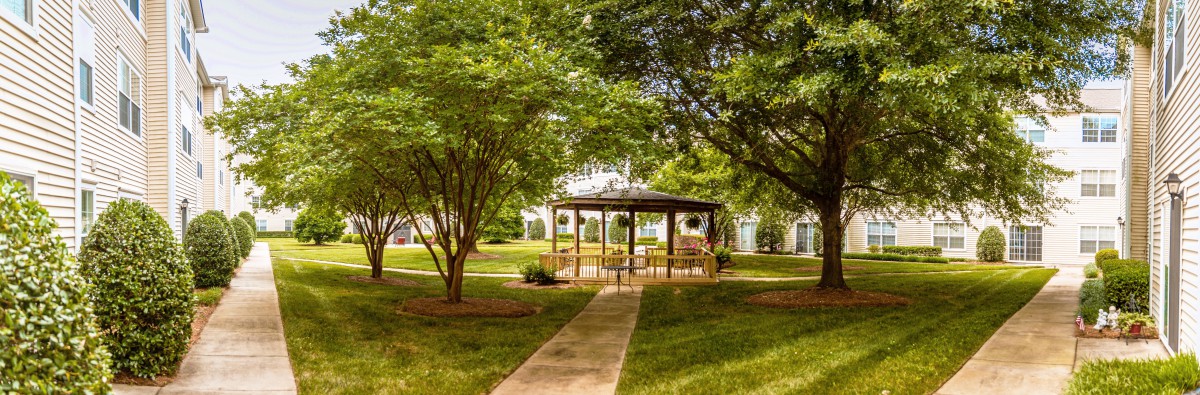 A peaceful park setting featuring a wooden gazebo surrounded by lush trees and greenery along a concrete pathway.