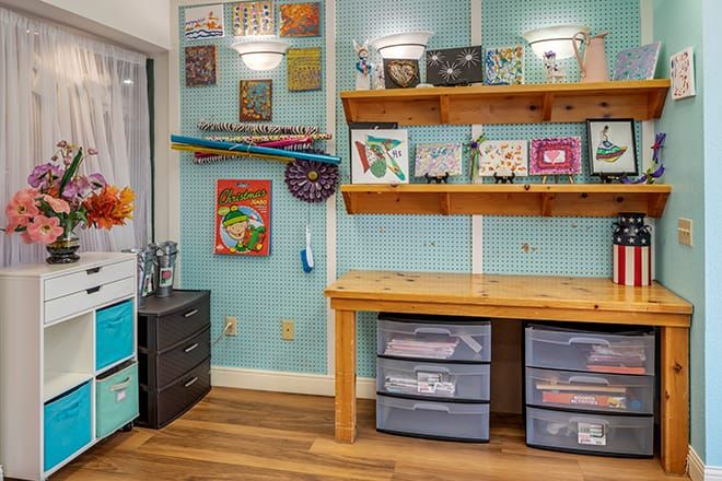 A well-organized craft room with a wooden work table, shelves displaying various art pieces, and storage bins for supplies.