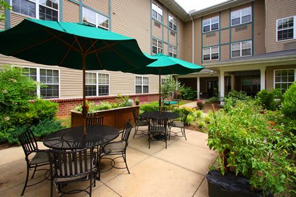 A cozy outdoor patio area with tables, chairs, and green umbrellas surrounded by lush greenery in a modern residential complex.
