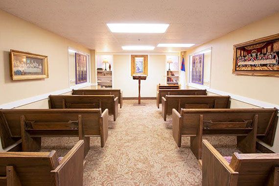 A small chapel interior with wooden benches and a central podium, illuminated by overhead lights.