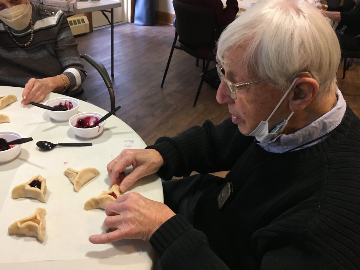 An elderly man is sitting indoors at a table, wearing a mask and engaging in a crafting activity.