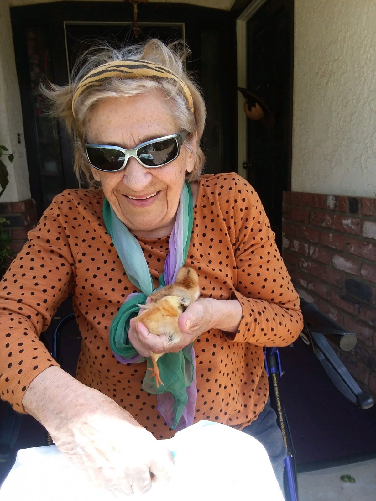 An elderly woman wearing sunglasses and a scarf, smiling while holding a small chick outdoors.