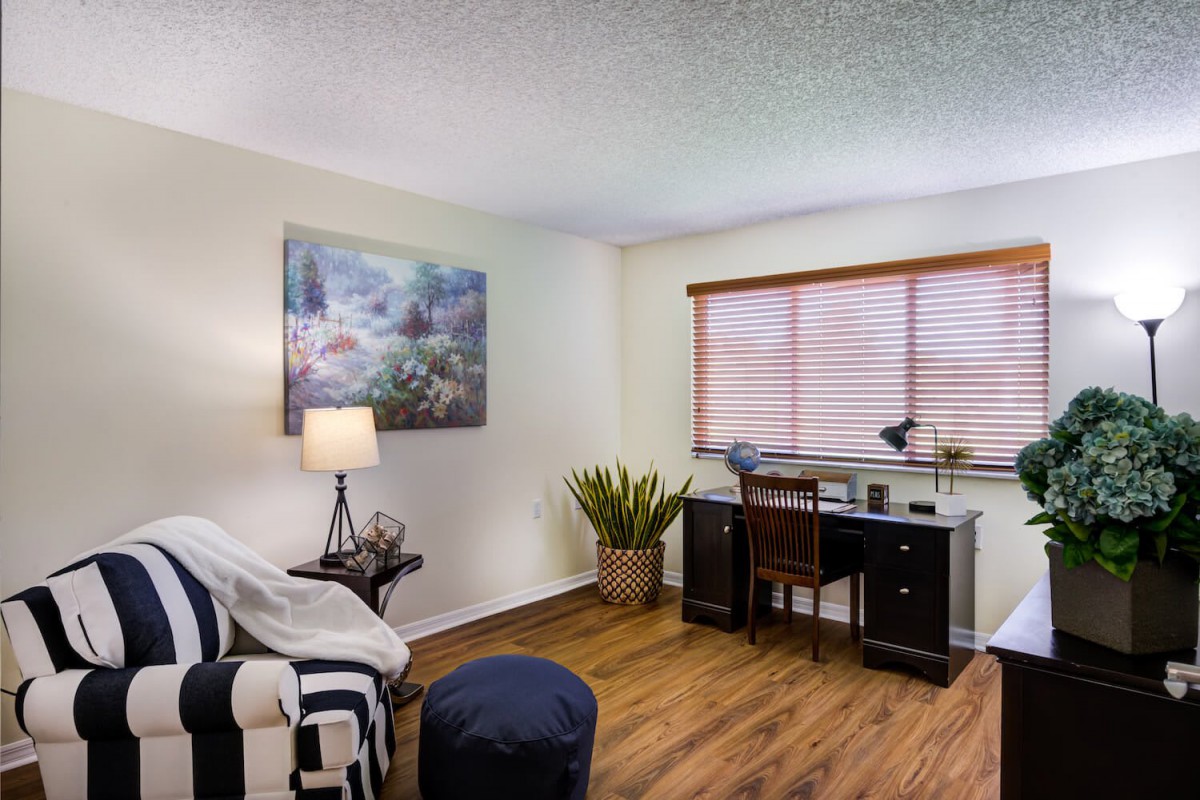 A cozy home office with a desk, chair, and lamp beside a window with blinds. A plant and painting add decor to the room, which features wood flooring.