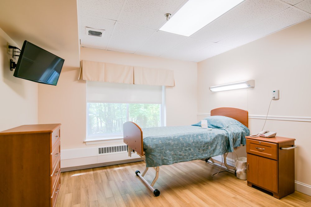A well-lit hospital room featuring a bed, a television, and light wooden furniture.