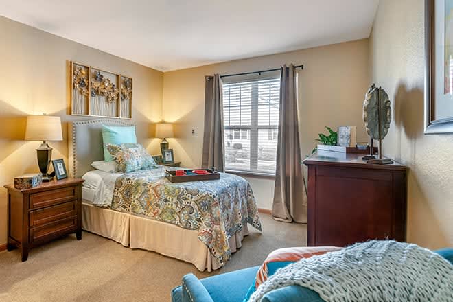 A cozy bedroom with a single bed, decorated with a floral quilt and cushions. The room features a large window with curtains, a lamp on a bedside table, and various decorative items.