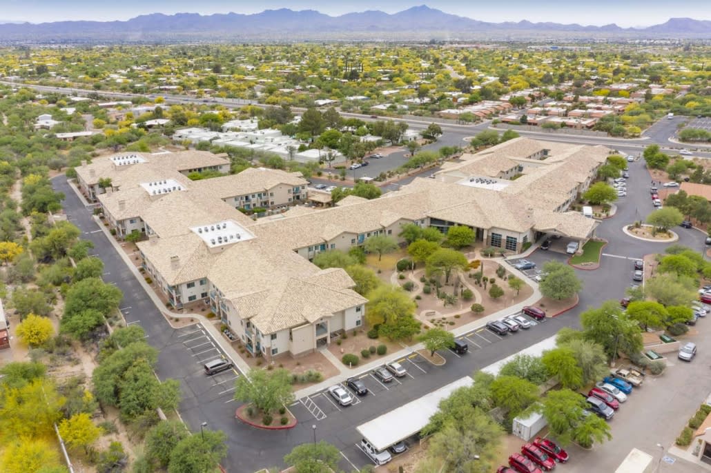 An aerial view of a residential complex with parking areas, surrounded by trees and set against a mountainous desert backdrop.