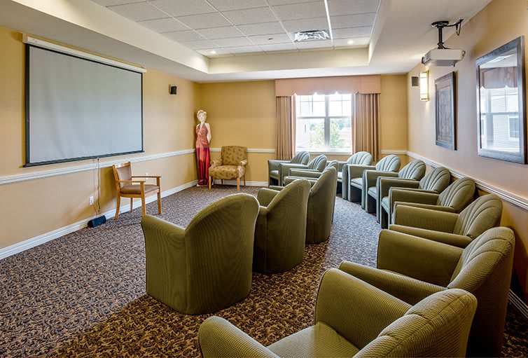A cozy meeting room setup with comfortable chairs facing a screen, carpet flooring, and natural light from a window.