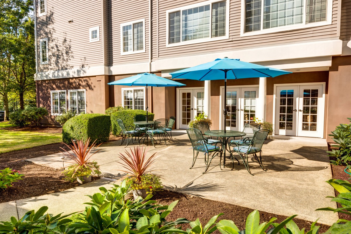 A charming outdoor patio with tables and blue umbrellas, surrounded by lush landscaping in front of a residential building.