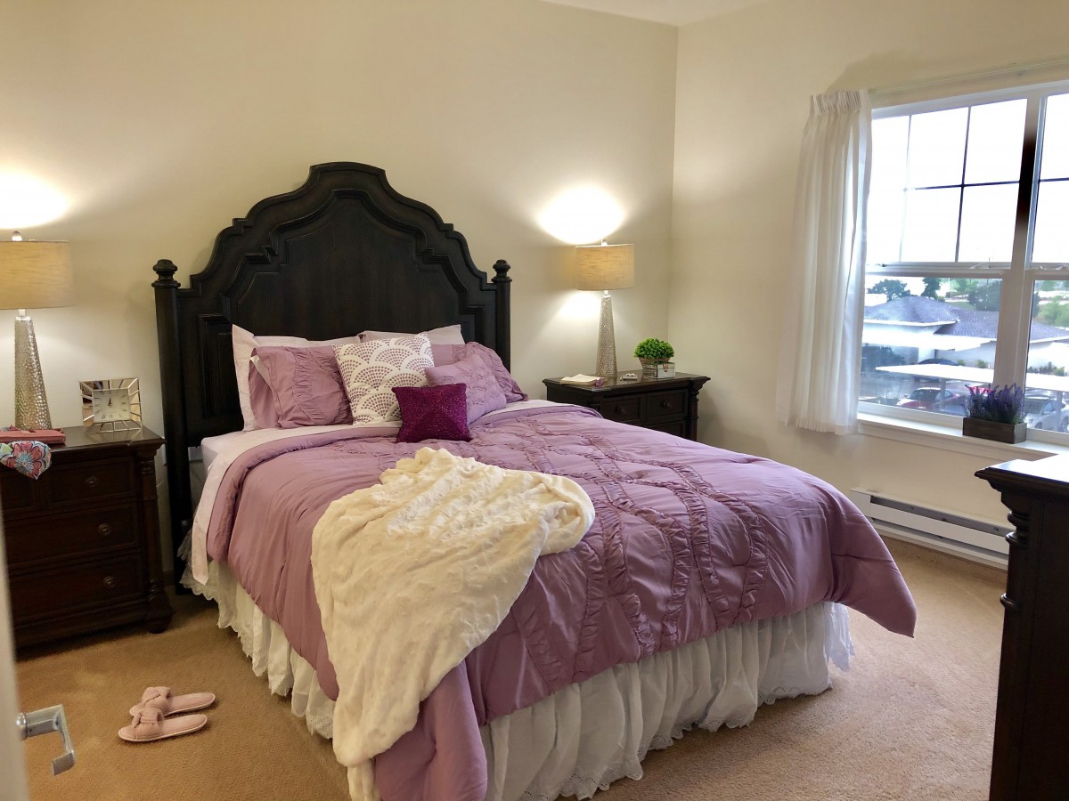 A cozy bedroom with a purple-themed bedspread and decorative pillows, softly lit by a lamp on the nightstand.