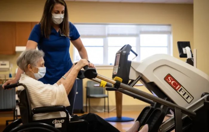 A healthcare professional assists an elderly person in a wheelchair with exercise equipment in a rehabilitation setting, both wearing masks.