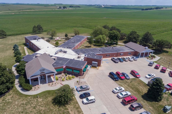 Aerial view of a large building with surrounding fields and a parking lot filled with cars.