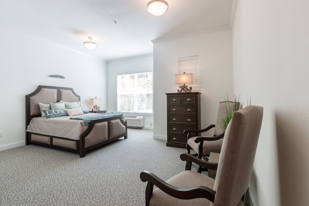 A cozy bedroom with a bed, dresser, and chairs by the window.