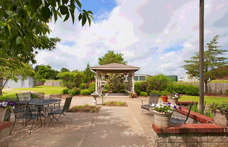A serene garden setting featuring a central gazebo surrounded by patio chairs and lush greenery.