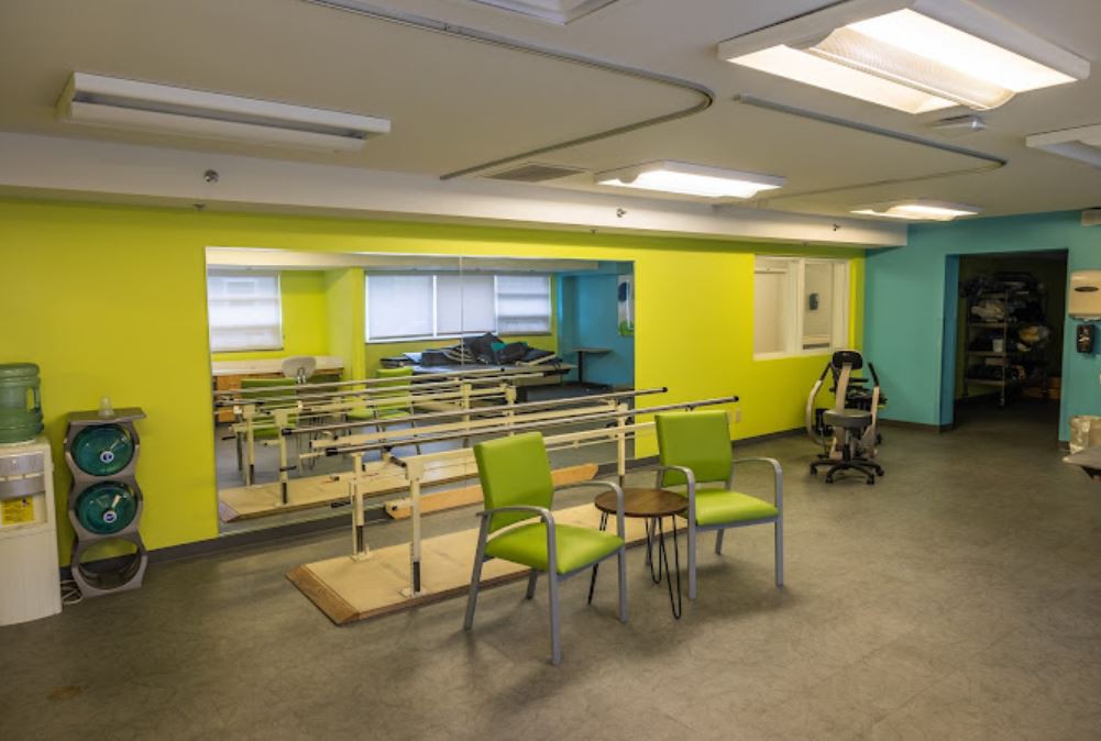 A brightly colored rehabilitation room equipped with parallel bars, a mirror, and chairs for physical therapy.