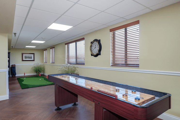 A spacious game room featuring a shuffleboard table, large windows, and a wall clock.