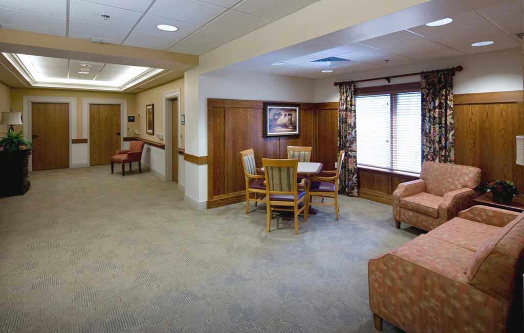 A comfortable lounge area with chairs and a table set on a carpeted floor, featuring large windows and decorative elements.
