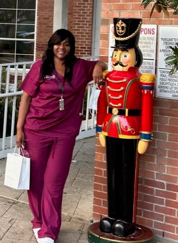 A person in scrubs poses next to a large decorative nutcracker statue outdoors.