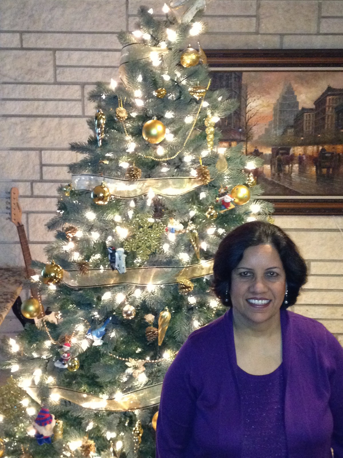 A woman smiling in front of a decorated Christmas tree adorned with lights and ornaments inside a cozy home.