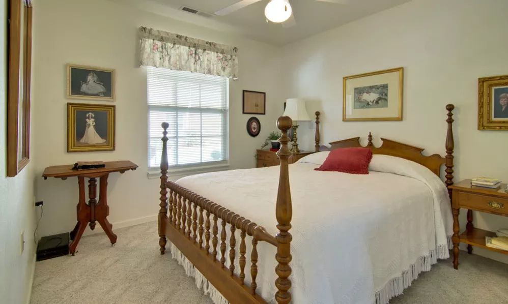A cozy traditional bedroom featuring a wooden bed frame and white bedding, complemented by wall art and a lamp near the window.