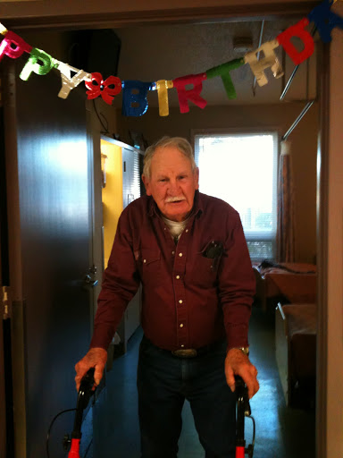 An elderly man using a walker stands happily in front of birthday decorations, celebrating a special occasion.