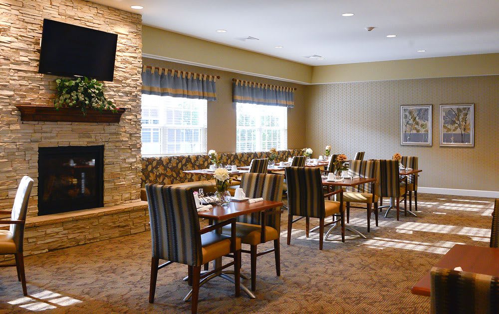 A cozy dining area with tables and chairs, featuring a fireplace and natural light from large windows.