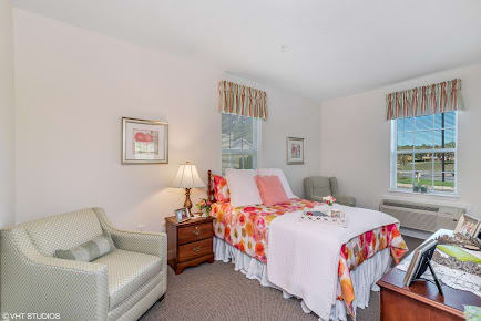 A cozy bedroom interior with a bed adorned in floral bedding, a lamp on a nightstand, and comfortable seating by the window.