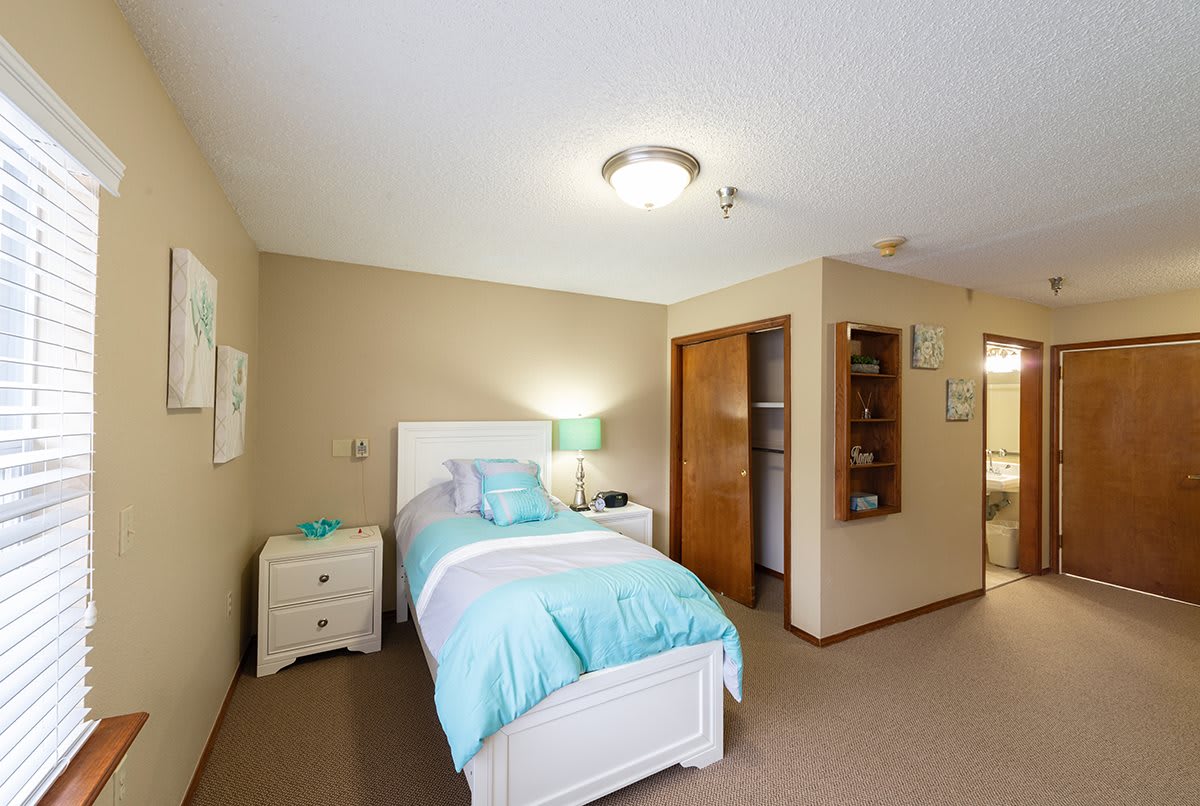 A cozy bedroom with a single bed featuring turquoise bedding, a nightstand with a lamp, and an open closet.