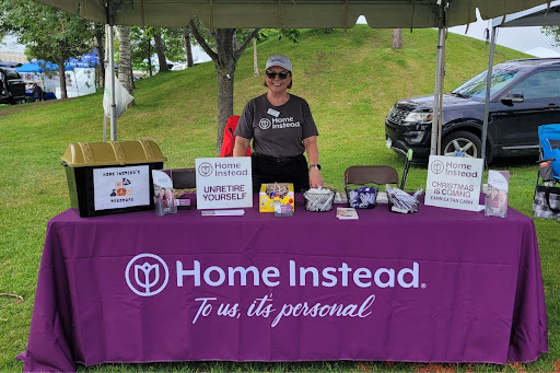 A volunteer stands at a Home Instead booth during a community event, providing information and resources.