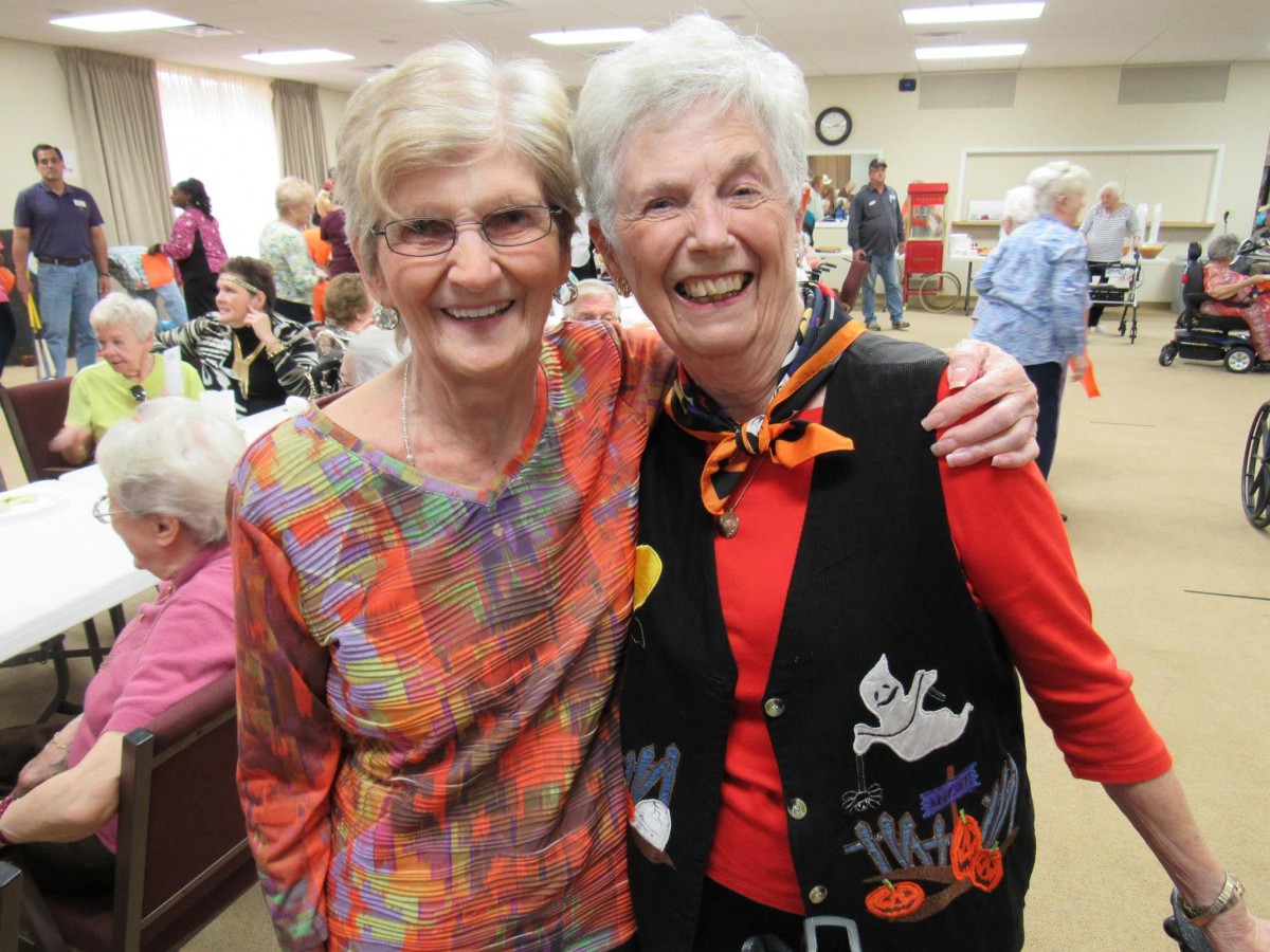 Two elderly women smile warmly for the camera at a community event, dressed in colorful and festive attire.