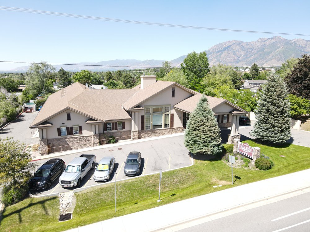 Aerial view of a large house with multiple cars parked in the driveway, surrounded by trees and mountains in the background.